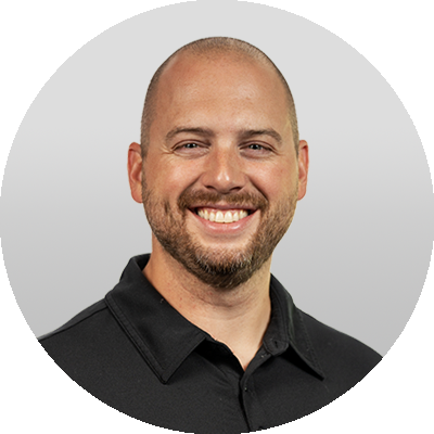 person smiling, wearing black polo shirt, shaved head with beard, light gray gradient background