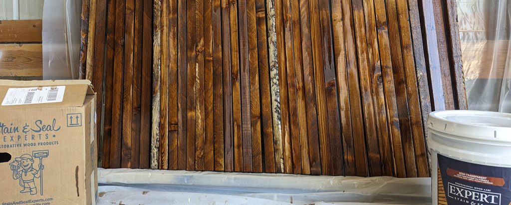 Stacked wood panels with a cardboard box and a white container labeled "Expert Stain & Seal" in the foreground.