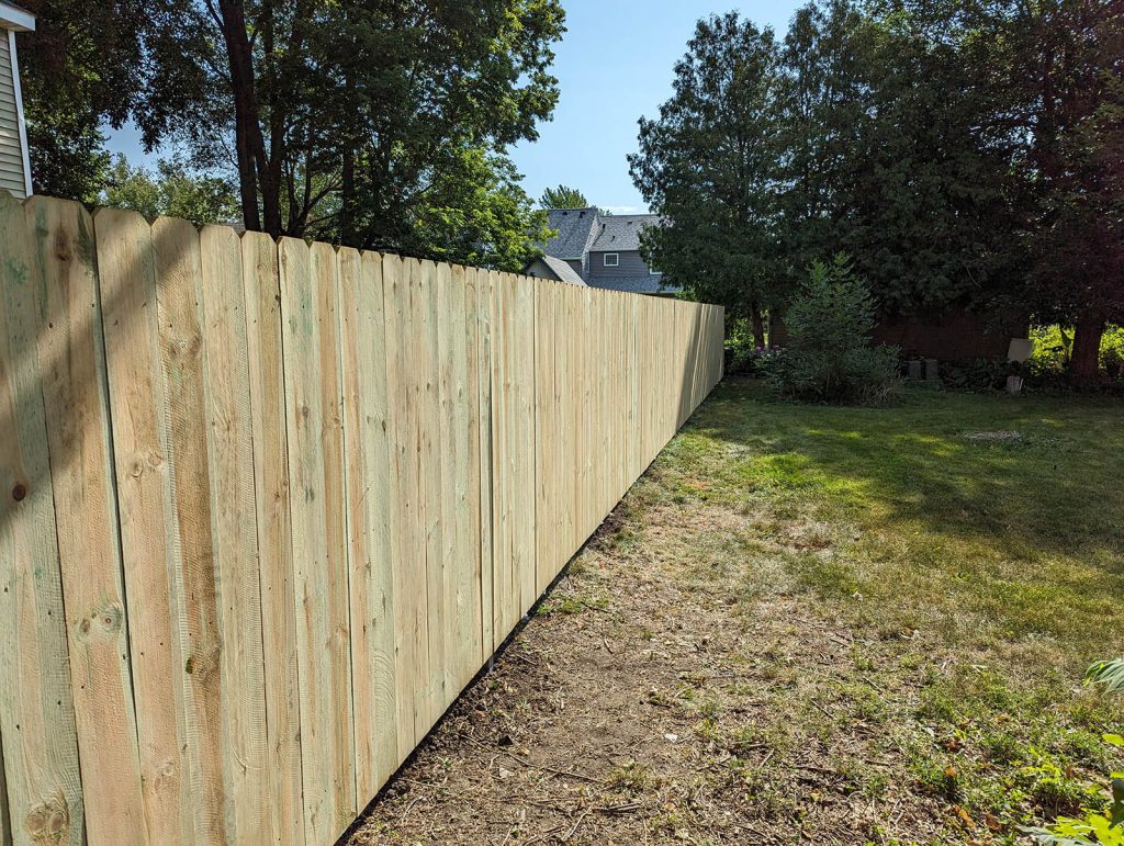 Long wooden fence along a grassy backyard with trees and houses visible in the background.