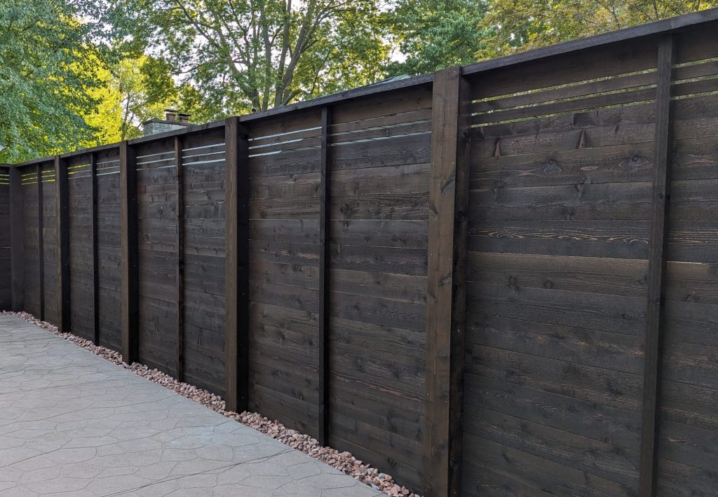 Tall stained wooden privacy fence with slats near top, beside light stone walkway and outdoor trees.