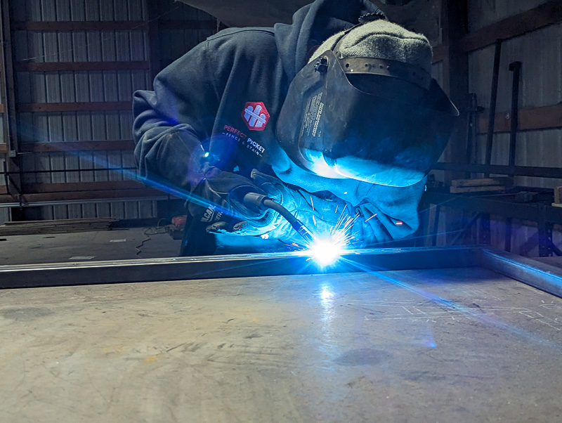 A welder working on a metal gate frame a residential wooden fence project in Waterloo, Iowa.