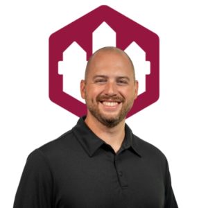 Man with a bald head and beard, wearing black polo, smiling in front of maroon hexagon logo.