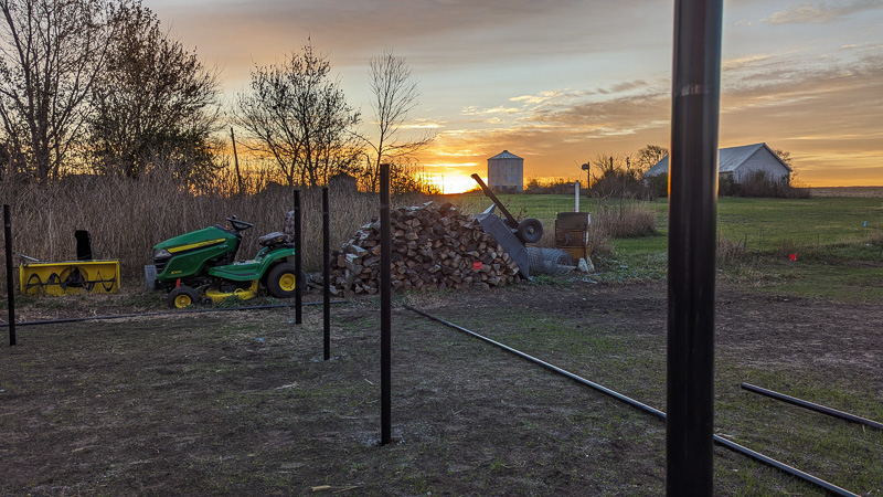 Rural scene at sunset with lawnmower, firewood, wood splitter, barn, silo, and bare trees.