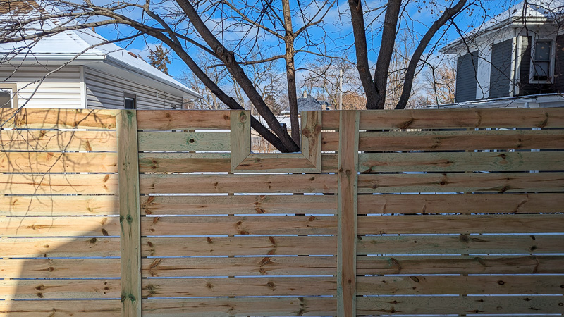 Wooden fence with cutout for tree trunk, situated in an outdoor residential area on a sunny day