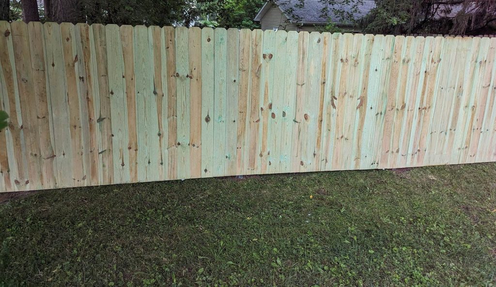 Newly constructed tall wooden fence with scalloped top edge on a grassy area, trees and house in background.