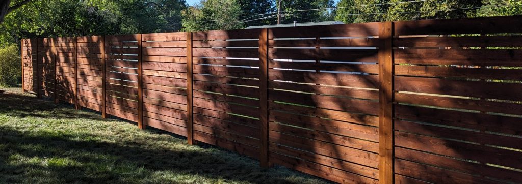 Wooden horizontal slat fence with gaps in outdoor area with grass and trees.