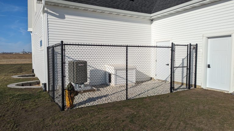 Black fence encloses air conditioning unit on gravel beside white building with a keypad-locked door.