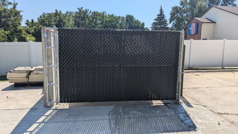 Chain-link gate with slats, pallet of materials, and building with "Motel 6" sign in outdoor area.