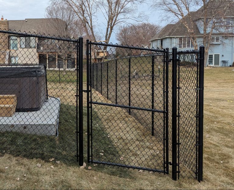 Chain-link fence with gate enclosing a backyard hot tub on a concrete slab, overcast sky, houses nearby.