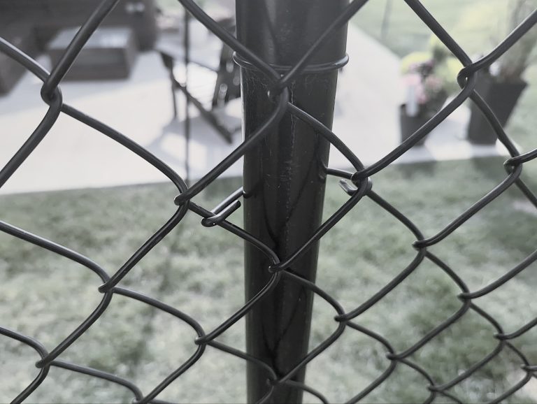 Close-up of a black chain-link fence with a metal post, patio furniture, plants, and grass in background.