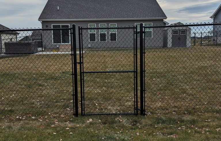 Black chain-link fence and gate in front of a grey house with a shed and hot tub in the backyard.