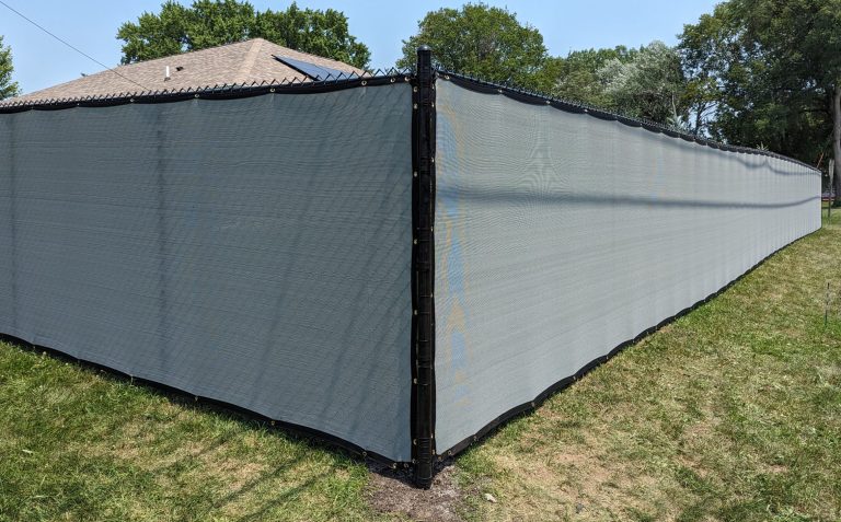 Gray mesh screen on chain-link fence, creating privacy on grassy lawn; house and trees visible.