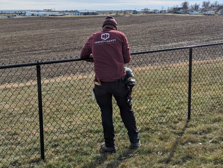 Person in maroon jacket working on chain-link fence beside large tilled field and buildings.