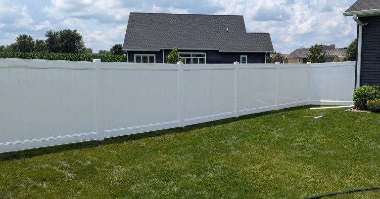 White vinyl privacy fence around a green lawn with residential houses and a blue sky in the background